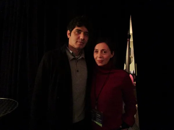 A dark-haired white man wearing a black coat and grey shirt stands with his arm around a brown-haired white woman in a cherry red pullover, in front of black velvet curtains.