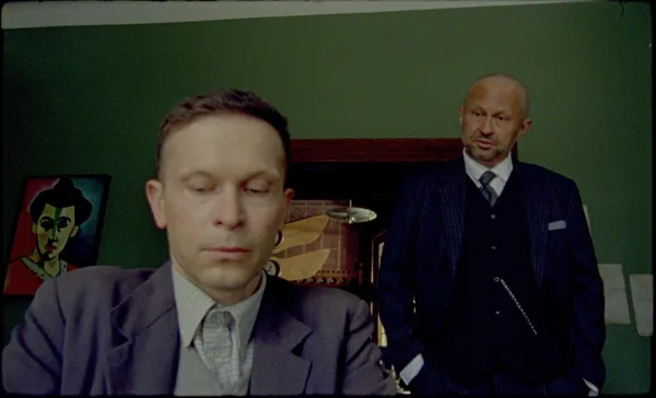 A white man sits down in the foreground while another, slightly older and wearing a rather striking blue suit with a silver watch chain, observes him from behind. in a room with paintings on the walls