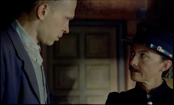 Two white men stand in front of a wooden door; the taller, younger one is looking down at the uniformed postmaster.
