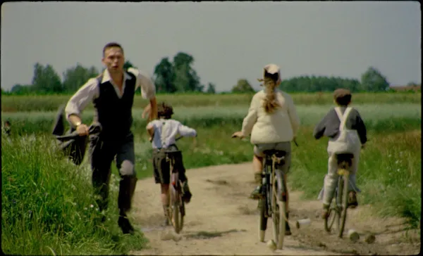 A still from the film The Assistant. A white man in a shirt and waistcoat, carrying a jacket, runs at full pelt down a country road as three children cycle in the opposite direction.