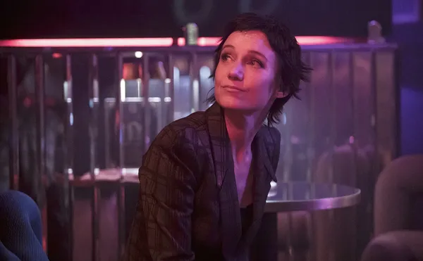 A still from the film Odyssey. A white woman with short dark hair sits in a nightclub illuminated by pink light.