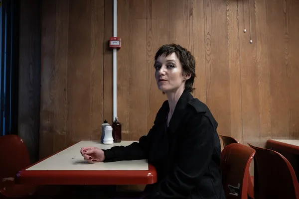 A still from the film Odyssey. A white woman with short dark hair, dressed in black, sits alone at a formica table in a café with wood veneer walls.