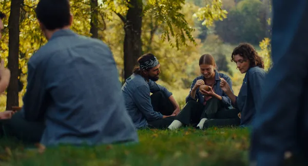 A still from the film Prime. A group of similarly clad young people sit among trees, smoking and eating food.