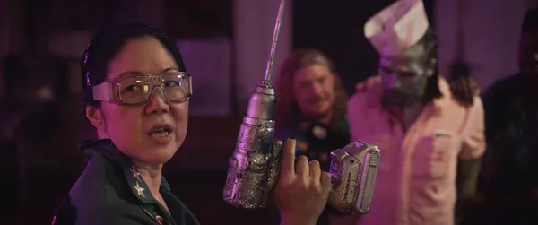 A still from the film Queens Of The Dead. An East Asian woman wearing goggles holds up a drill as a zombie in a chef's uniform is dragged towards her.