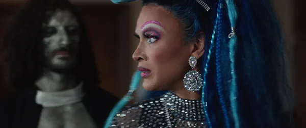 A still from the film Queens Of The Dead. A Black drag queen with fabulous long blue hair is approached by a dead-looking clergyman.