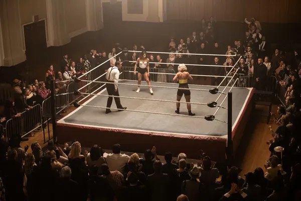 A still from the film Queen Of The Ring. Two white women face off in a wrestling ring in a large hall, surrounded by a standing audience.