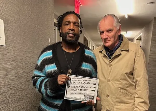 A middle-aged Black man in a stripey sky blue and black cardigan stands in a corridor beside an older white man in a long beige coat.