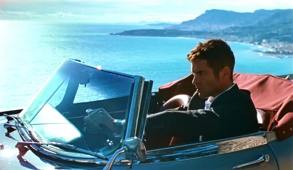 A still from the film Reflection In A Dead Diamond. A white man in a dark suit drives an open-top car along the Cote D'Azur coast road.