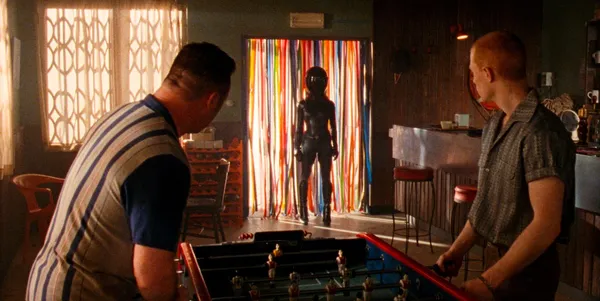 A still from the film Reflection In A Dead Diamond. Two white men playing table football in a quiet bar look towards the entrance as a woman in a black leather catsuit and black motorbike helmet enters.