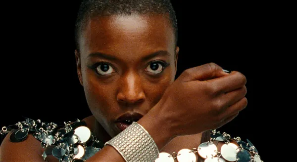 A still from the film Reflection In A Dead Diamond. A Black woman in a dress made from silver spangles speaks into a silver bracelet communicator.