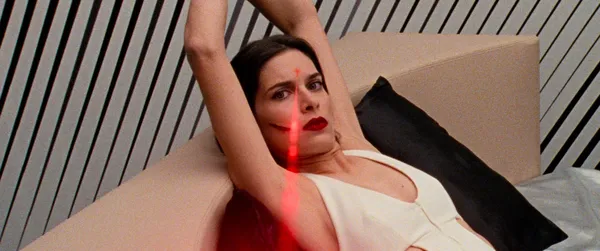 A still from the film Reflection In A Dead Diamond. A white woman wearing a white dress and vivid red lipstick is tied up in a room whose walls are painted with diagonal black and white striped. A thin red laser beam is pointing at her forehead.