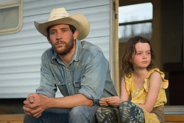 A white man wearing a short sleeved shirt and a cowboy hat sits on the steps of a trailer beside a white girl wearing a yellow frilly top.