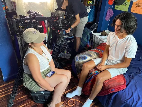 A behind the scenes shot from the film Remaining Native. A Haudenosaunee woman squats down to talk to a Paiute teenage boy as he sits on his bed. Filmmaking equipment is stacked up at the other side of his room.