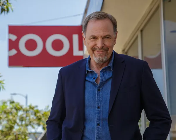 The actor and director Richard Green. A smiling, bearded white man stands outside a low building under a light blue sky, next to a sign which says 'cool'.