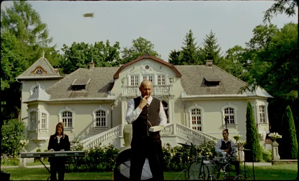 Two white women play the kayboard and drums in front of a large country house, as a middle aged white man stands in the foreground looking at a book.