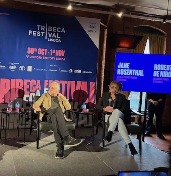 A older white man and woman sit in chairs on a stage surrounded by Tribeca festival banners.
