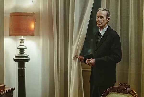 An older white man stands by a tall window at night, the curtain in his hand.