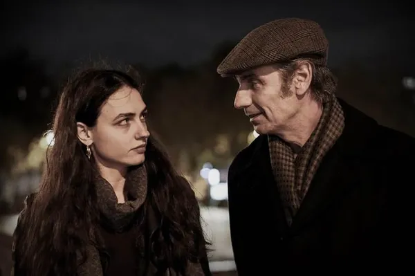 A young dark-haired white woman in a warm dark coat and a middle-aged white man in a brown coat and a flat cap talk to one another in an urban street at night.
