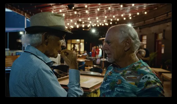 Two older white men - one wearing a blue polka dot shirt with a hat, the other a tropical-themed T-shirt - face one another in a bar, looking thoughtful.