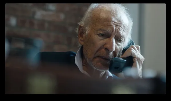 An older white man speaks into the receiver of a rotary dial telephone.