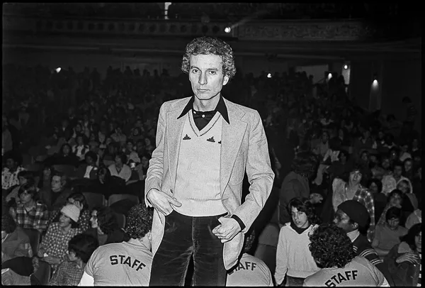 A black and white image of a white man wearing a v-neck sweater and velvet trousers, standing on a stage in front of a crowd.