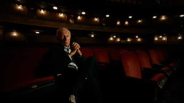 A smartly dressed older white man sits on a red velvet theatre seat in an otherwise empty auditorium.