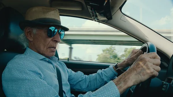 An older man wearing a blue patterned shirt, a hat and blue-tinted mirror-shades, drives a car.