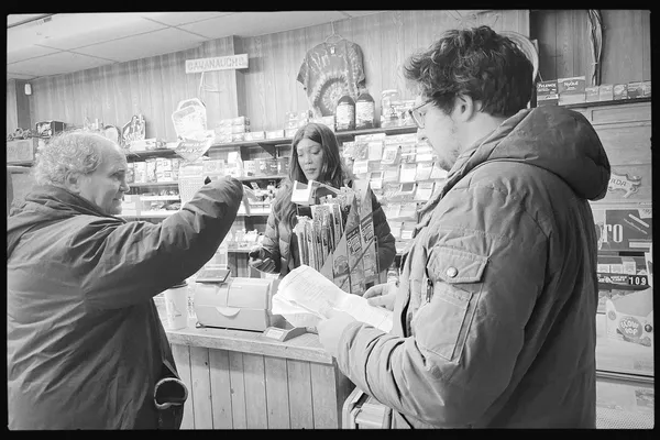 A behind the scenes shot from the film Stationed At Home. Two white men and a Black woman prepare to shoot a scene in a convenience store. The woman is behind the counter and one of the men is holding a script.