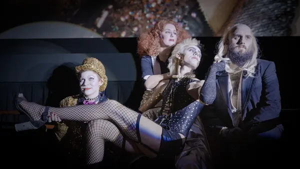 A still from the film Sane Inside Insanity: The Phenomenon Of Rocky Horror. A Rocky Horror shadow cast performs Sweet Transvestite, with a white transvestite in a basque and fishnet stockings lounging sideways in a chair.