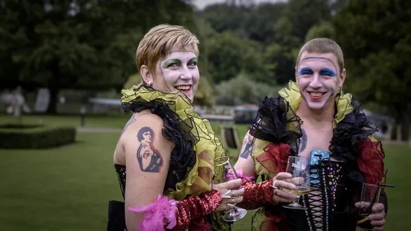 A still from the film Sane Inside Insanity: The Phenomenon Of Rocky Horror. Two white people in fabulous feather boas stand side by side in a large garden.