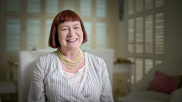 A still from the film Sane Inside Insanity: The Phenomenon Of Rocky Horror. A middle aged white woman with bobbed red hair sits on a white chair in a white room.