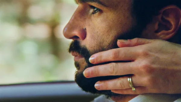 A still from the film Swimming Home. A pale hand rests across the neck of a white man with dar, curly hair and beard as he sits in a car.