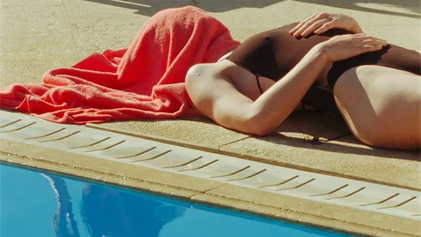 A still from the film Swimming Home. A white woman in a brown swimsuit lies beside an outdoor swimming pool, with an orange towel over her head.