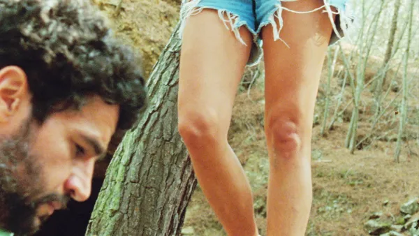 A still from the film Swimming Home. In woodland, a white man with curly dark hair and beard rests his head close to the camera. We can also wee the slender legs of a white woman, in cut-off denim shorts.
