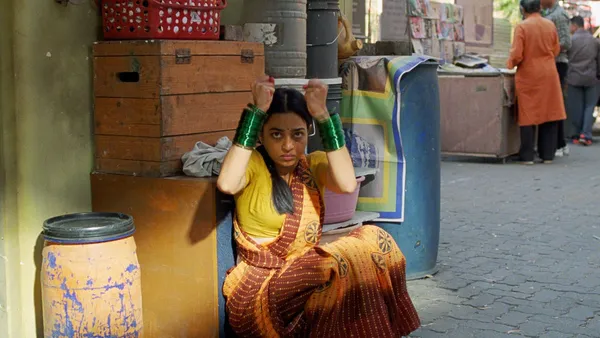 A still from the film Sister Midnight. A young Indian woman squats down beside some boxes at the edge of a market square. Her wrists are covered in shimmering green bangles.