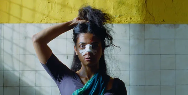 A still from the film Sister Midnight. A wide-eyed young Indian woman with bandages across her nose stands in a tiled room holding her long hair up above her head.