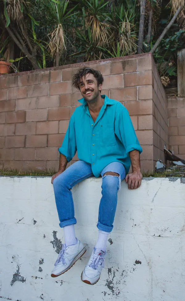 Sam Hayes. A man with light brown skin, a moustache and tousled hair sits on a white painted wall above which palm trees are growing. He's wearing ripped blue denim jeans and a light blue shirt.