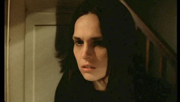 A still from the film Weightless. A dark-haired young white woman beside a door, with a staircase behind her.
