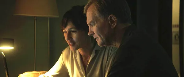 A still from the film Dead Language. A white woman and man sit side by side on a bed, talking unhappily.