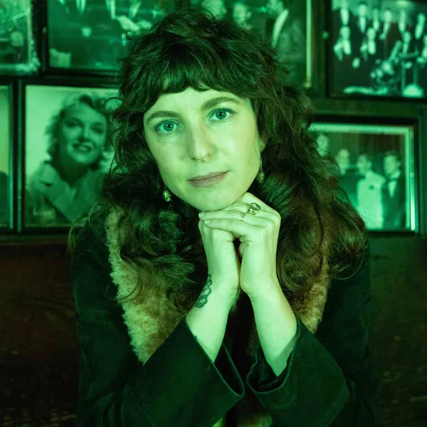 Portrait shot of Sasha Wortzel, it has been taken in a green light. She has shoulder length curly brown hair and is resting her chin on her hands. She is wearing a nose ring