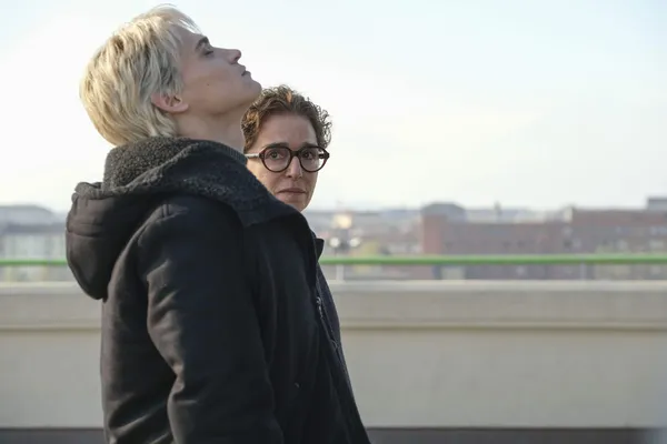 A still from the film La Gioia. A blond youngwhite man wearing a big dark coat tips back his head as he walks across an urban bridge beside a dark-haired, bespectacled middle-aged woman.