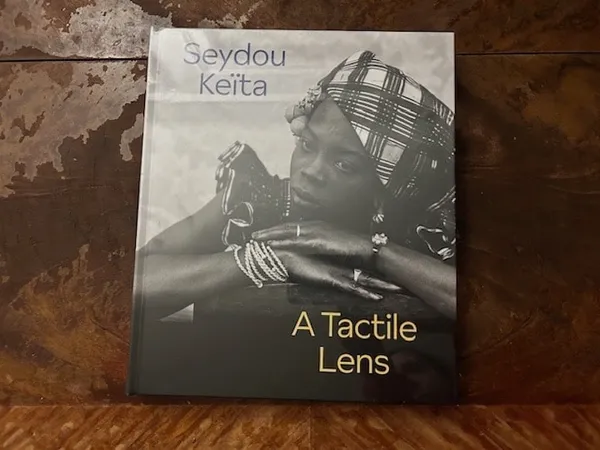 A book resting on a wooden surface. The cover is a black and white photo of an African woman. In the top left hand corner is the name Seydou Keïta; in the lower right hand corner the words 'A Tactile Lens'.