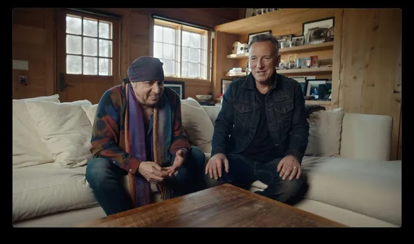 Two older white men - one dressed in colourful clothing with a scarf around his head, and one in denims - sit on a white leather corner sofa in a wooden cabin.