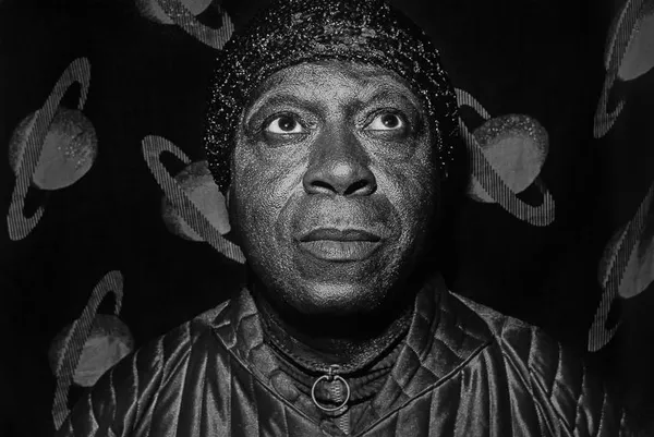 A black and white photo of a middle-aged black man wearing a beaded gap, against a background studded with ringed planets.