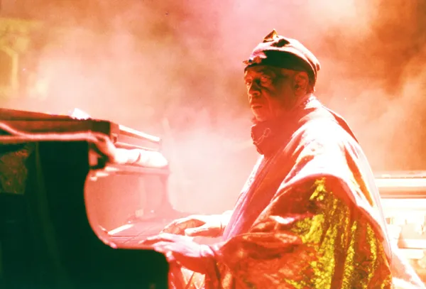 Against a background of orange-lit smoke, a middle-aged Black man dressed in diaphanous orange robes plays the piano.