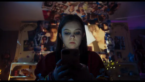 A still from the film Sweetness. A white teenage girl focuses on her mobile phone in a room full of posters and pictures of the same young white man.