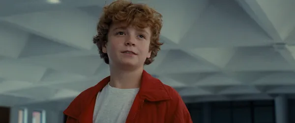 A still from the film The Plague. A white boy in his mean teens with wavy golden hair cynically surveys a room.