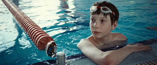 A still from the film The Plague. A white teenage boy with goggles push back over his short brown hair looks around as he holds onto the side of a swimming pool.