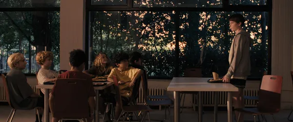 A still from the film The Plague. At night, a group of early teenage white boys sit around a table while another stands at a distance, behind another table, facing them like a nervous teacher.