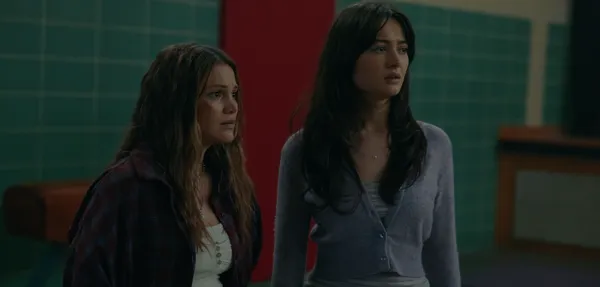 A still from the film This Is Not A Test. Two light-skinned young women with concerned expressions stand beside the turquoise tiled wall of a school gym.